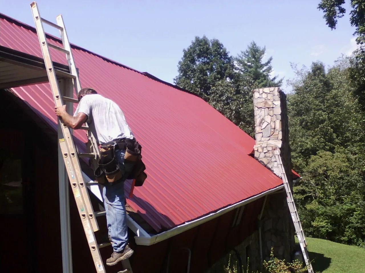 Roofer on a ladder beside a red metal roof and stone chimney — Soffit & Fascia Repair by Roofing Brothers in Sutton